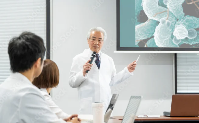 スクリーンを指しながら白衣姿の参加者に講義する講師