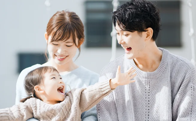 両手を広げて笑う子どもと、笑顔で見守る両親